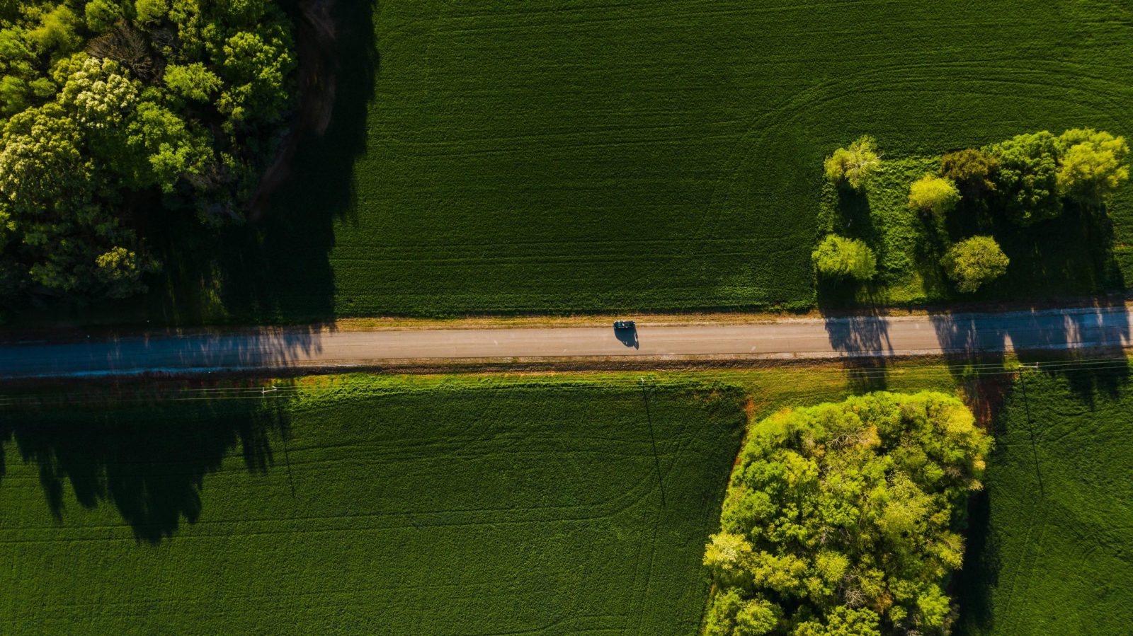 Aerial shot of a car driving on a tranquil road through lush green countryside on a sunny day.